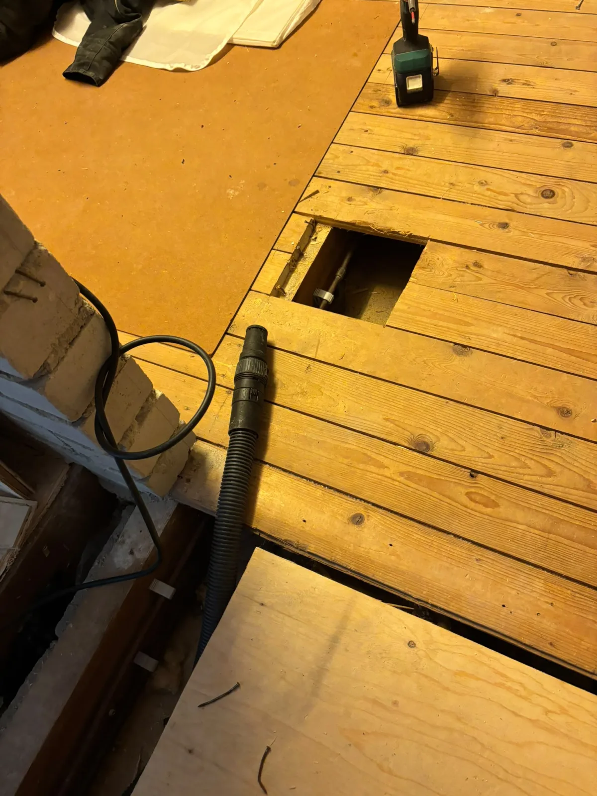 Houten vloer gedeeltelijk opengebroken, leidingwerk en ruimte eronder zichtbaar.